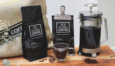 Fresh roasted coffee bags from Sea Island Coffee containing St Helena coffee beans, sitting on a wooden tray next to some flowers and a french press