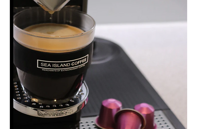 Sea Island Coffee cup with coffee being made from a Nespresso machine, next to some pink Nespresso compatible pods